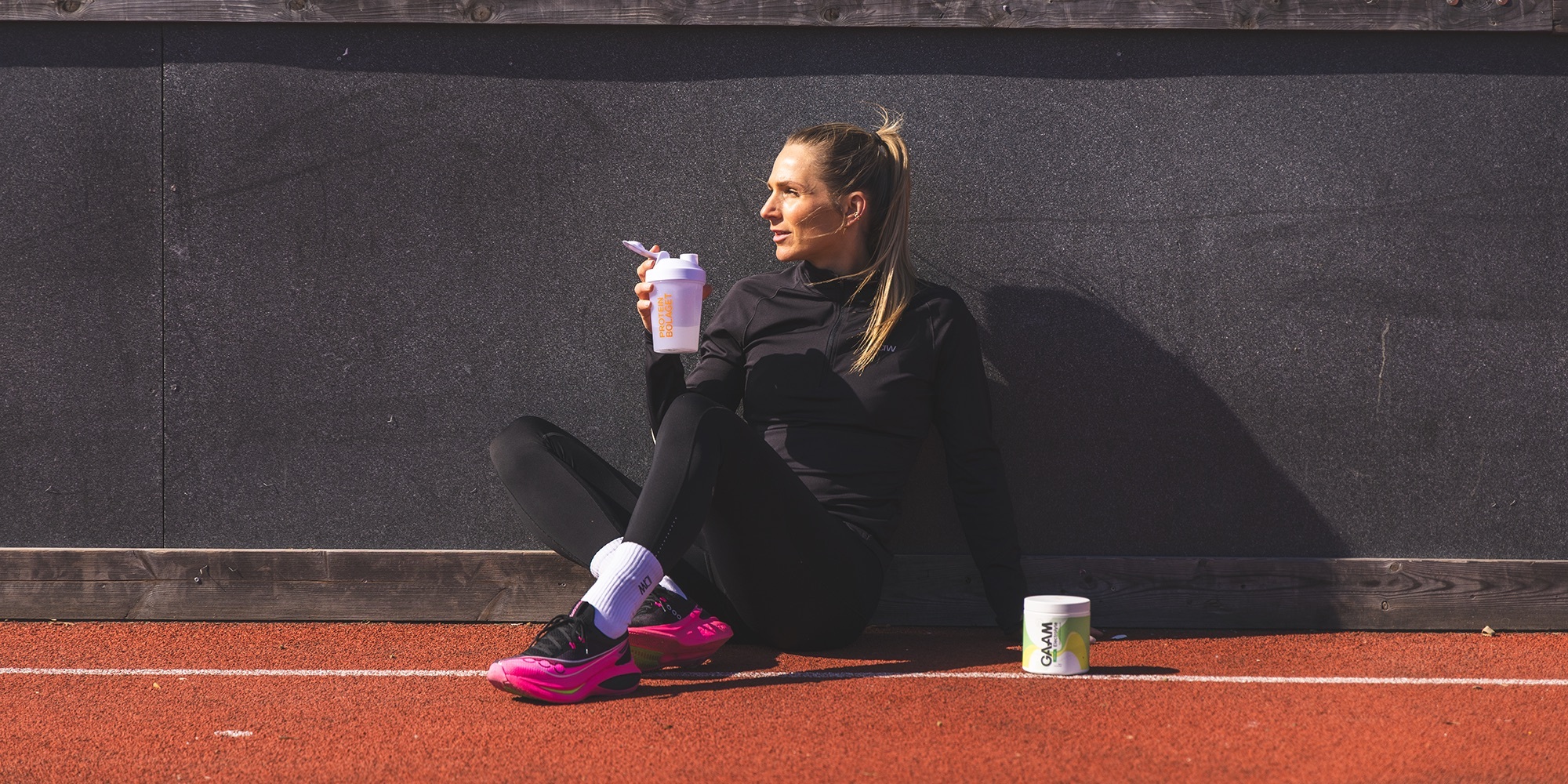 Kvinna sitter på löparbana efter löpning och dricker kosttillskott ur Proteinbolaget-shaker, med GAAM-burk bredvid.
