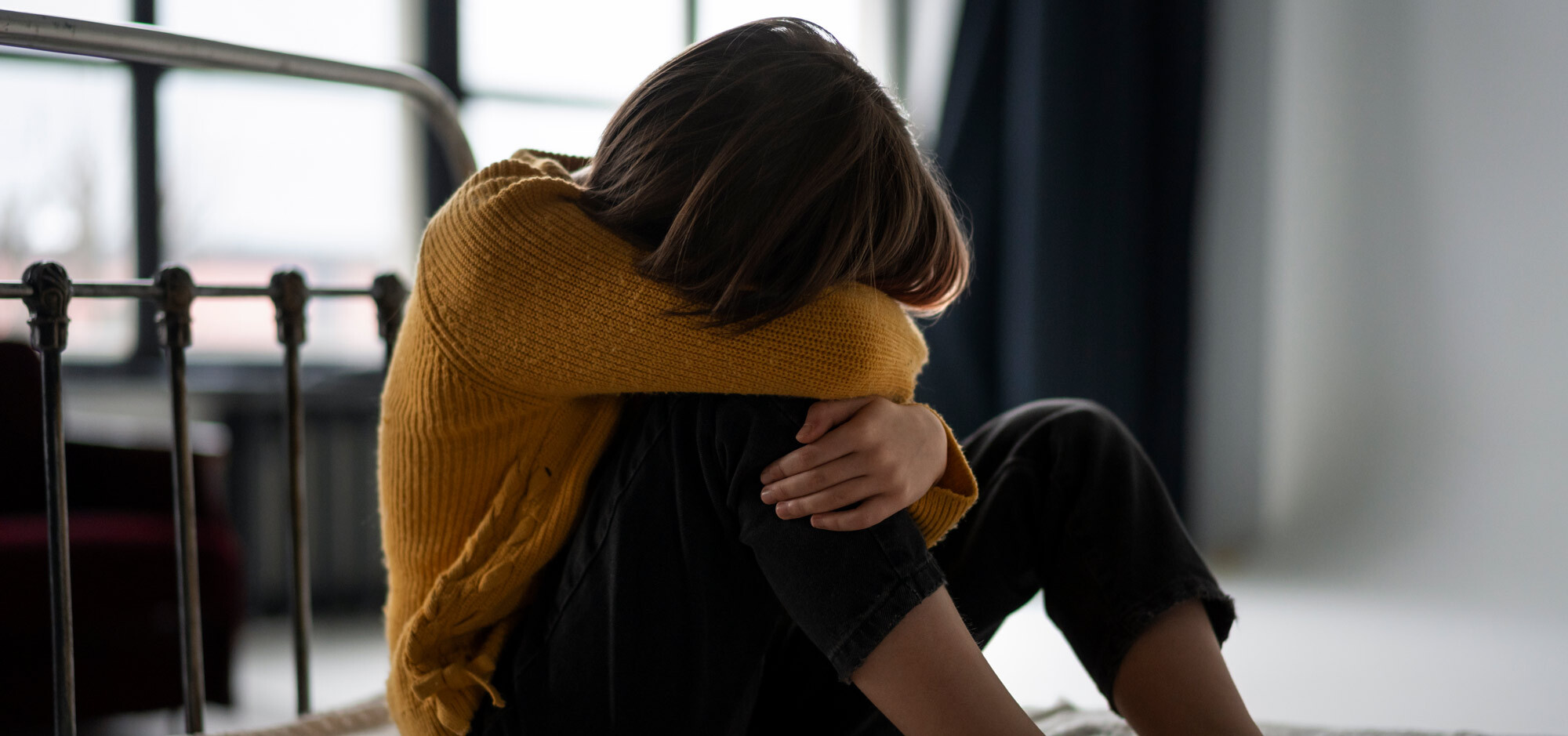 En person med gul stickad tröja sitter hopkrupen på en säng med armarna runt benen, i ett dunkelt rum – en bild som uttrycker djup trötthet och inåtvändhet, vilket kan spegla vardagen med ME/CFS.