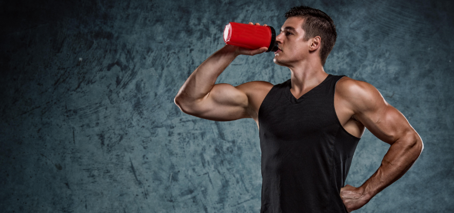 Muskulös man som står och dricker ur en röd proteinshake